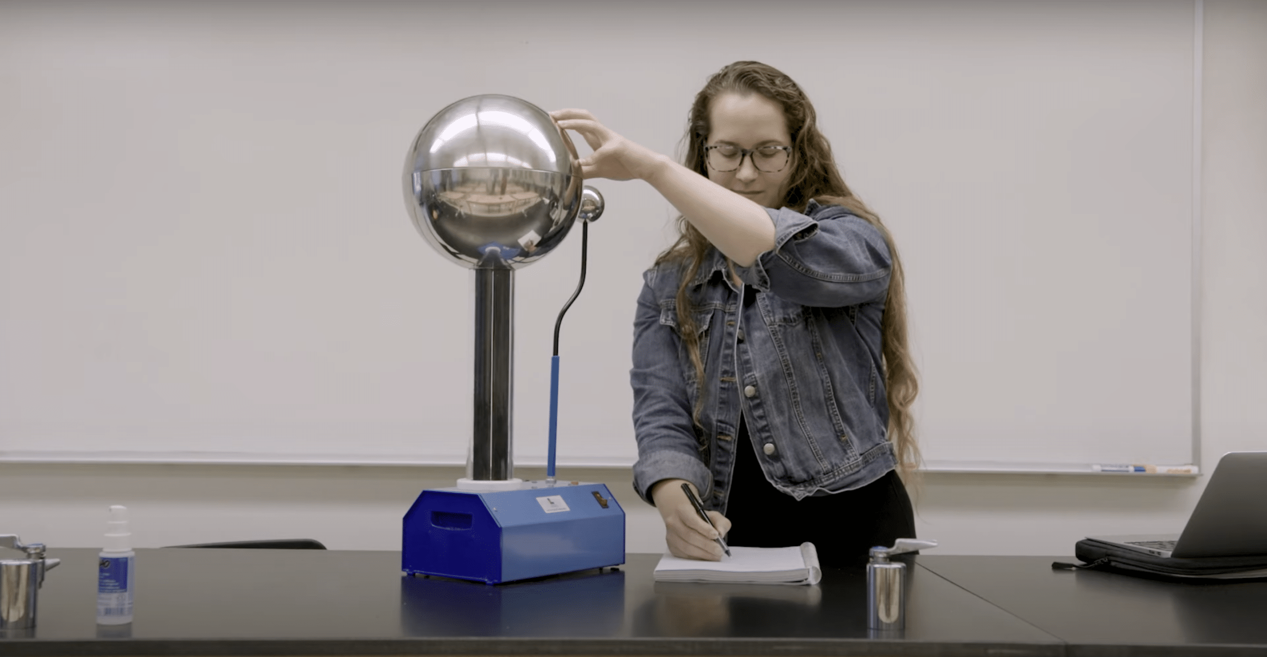 Young woman with scientific equipment touching a stainless steel ball and taking notes.